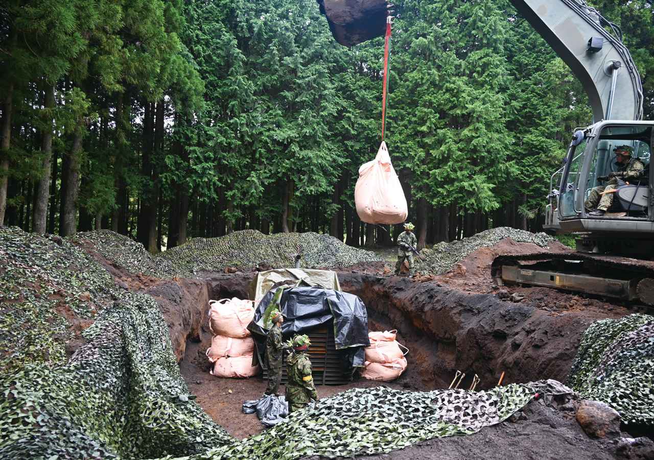 画像: ①重機を使用して指揮所を構築:10高射の指揮所を重機を使用して構築。油圧ショベルで陣地に土のうを積み上げて補強する