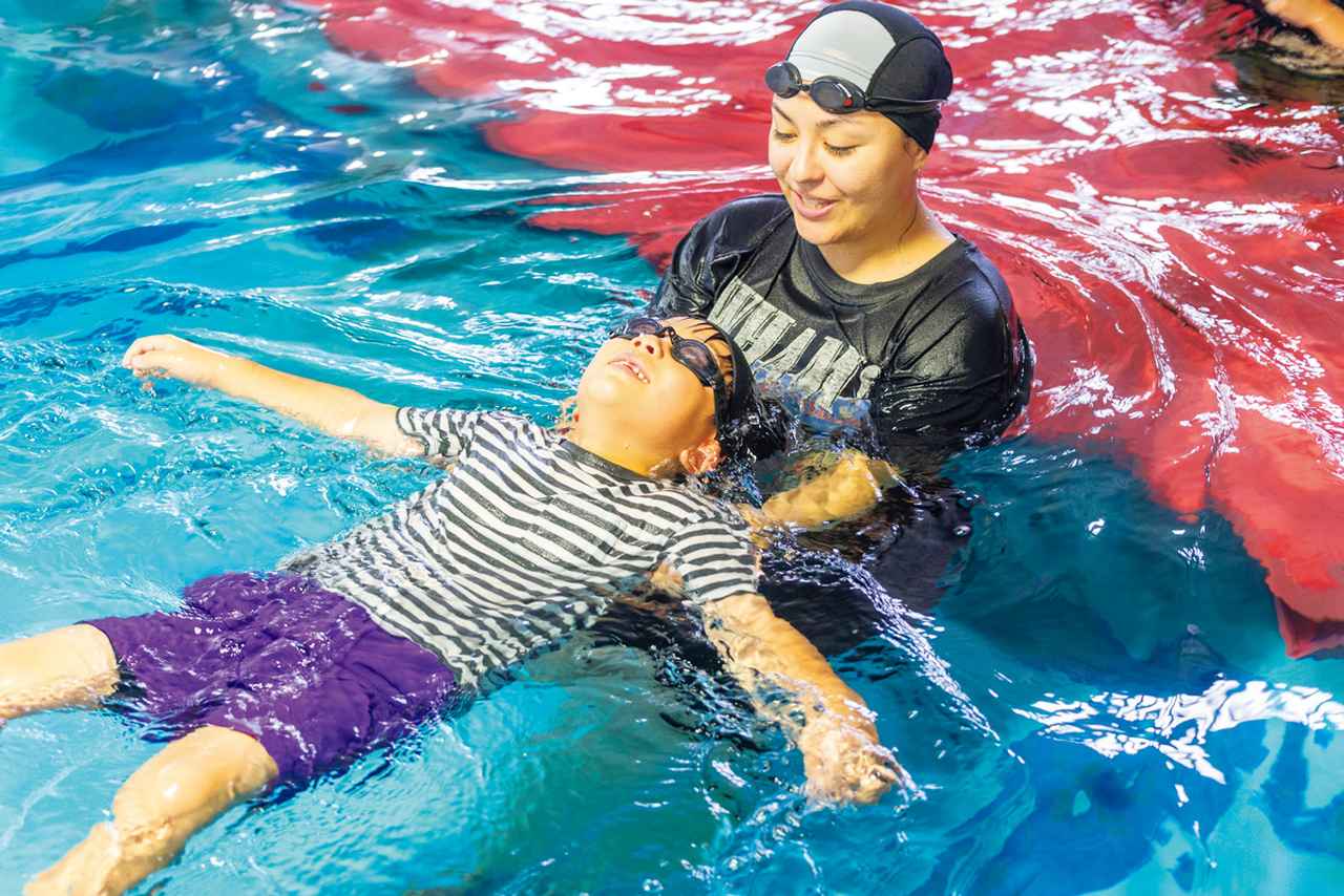 画像: 岡崎竜城スイミングクラブの「着衣泳」レッスンの1コマ。レッスン生の子どもが、服を着たままプールに入り、指導員に支えられながら浮く練習をしている