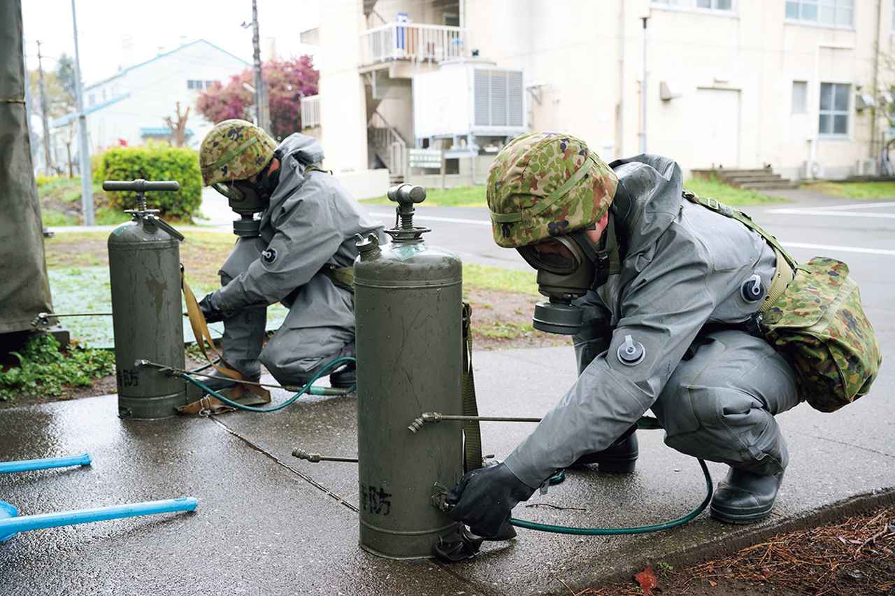 画像: 【除染器の使用前に除染剤を注入し加圧】携帯除染器を使用する前の準備をする受検者。除染剤を水で溶かして除染器に注入し、スプレーガンで噴霧することができるように、手動ポンプ(レバー)を押して加圧。防護服を着てのこの力作業は視界が悪く、まるでサウナの中にいるかのような暑さだという