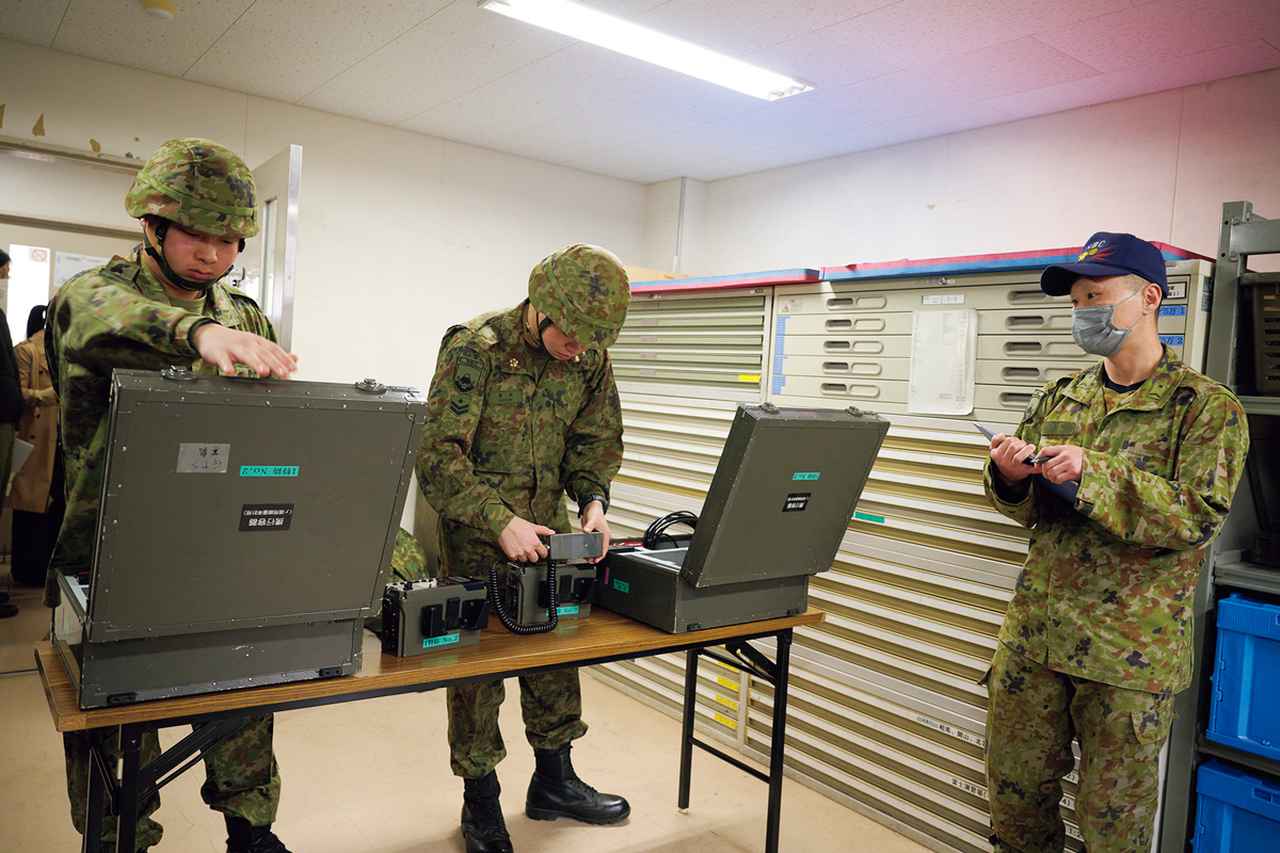 画像: 【検知器材の使用前の準備を実施】ガンマ線用線量率計の使用に関する実技検定を受ける受検者たち。専用のケースから取り出して組み立てるまでの工程を、いかに正確かつ迅速に行えるかが問われる。傍らに立つ検定官は「検定支援」の役割も担い、作業の間違いの指摘やアドバイスを行う