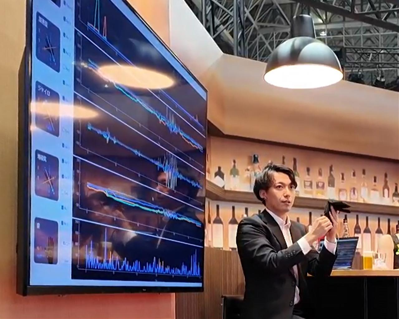 画像: ミニステージでデモを説明する万竝(まんなみ)亮（日立製作所 インダストリアルAIビジネスユニット）。センサー付きグローブは、コネクターなど小さな部品操作の感触や音も検知できます。